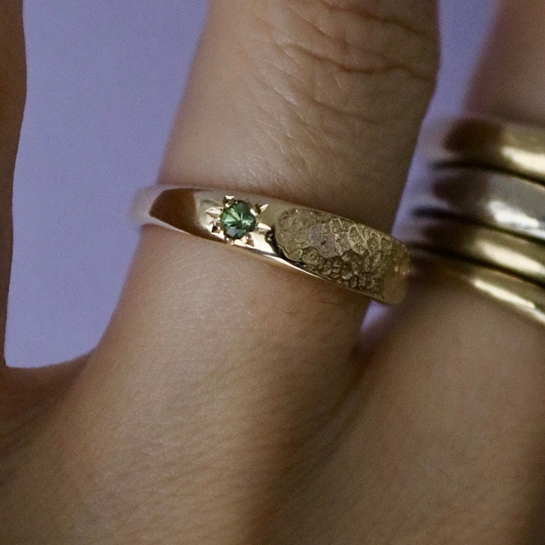 A polished yellow gold ring with a green star set tourmalime. The ring is on the wedding ring finger and is 5mm high. The nose print texture is on the face of the ring across the finger.
