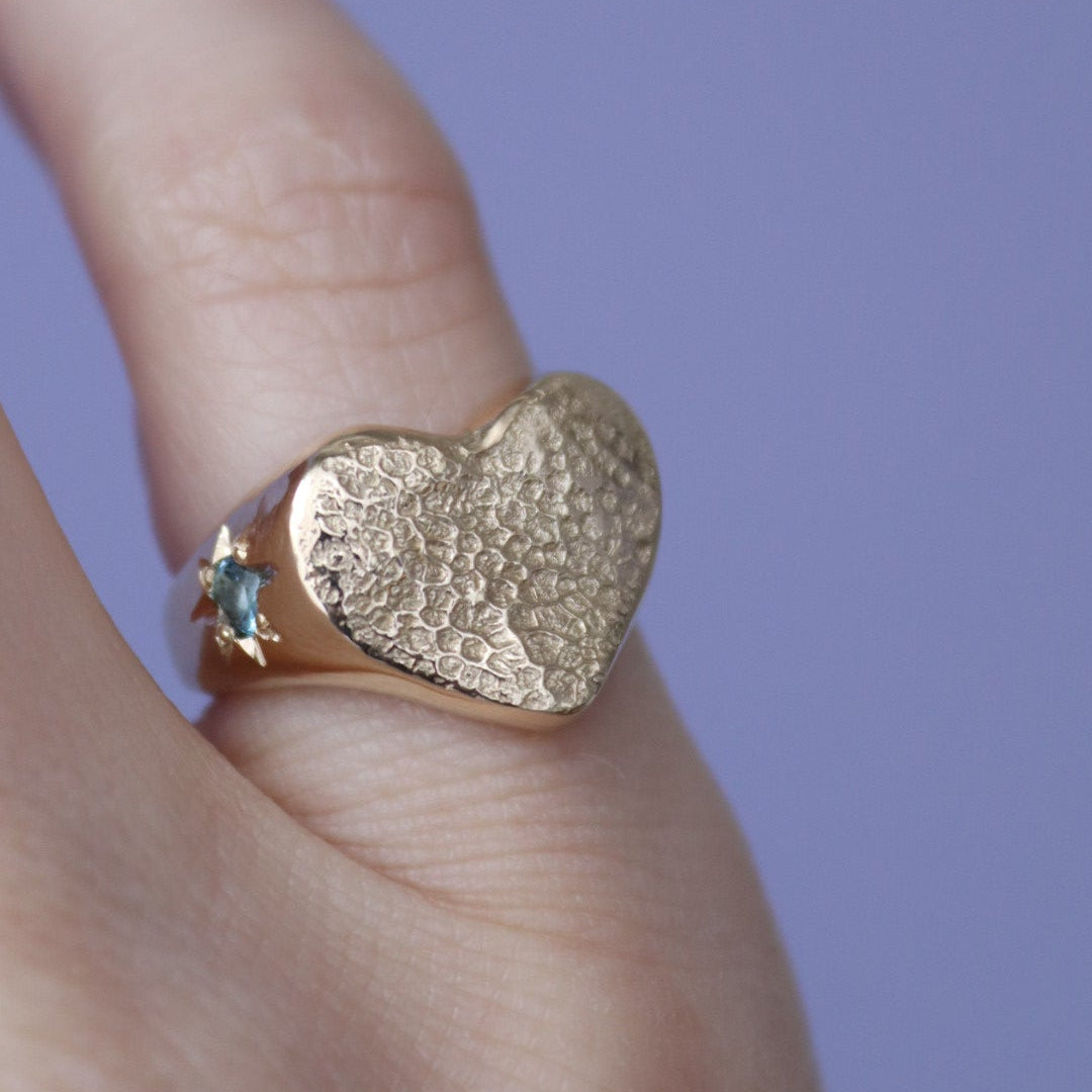 Gold heart-shaped ring with a blue gemstone on a finger against a purple background