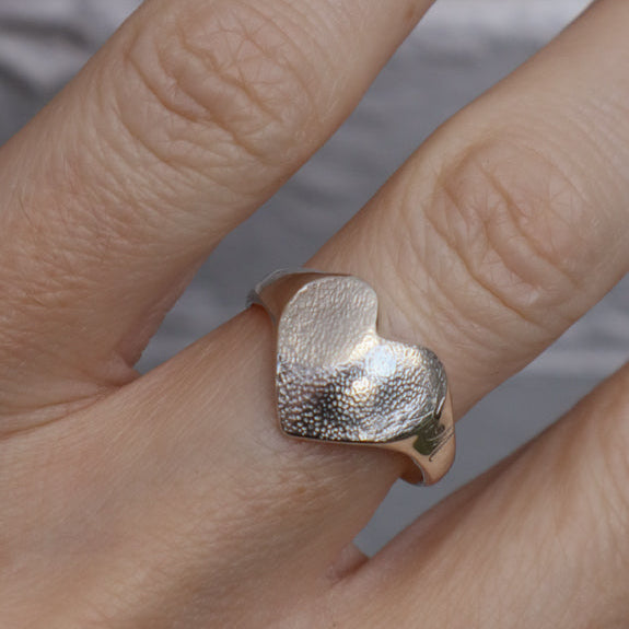 A silver ring heart shaped ring with a dog nose print texture on it, shown worn on a person's finger against a soft gray background.