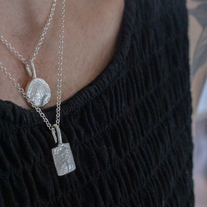 Close-up of a person wearing two sterling silver dog nose print pendants on a chain with round links. One rectangle one circular  