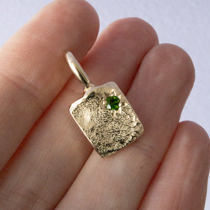 Gold rectangular dog nose print pendant with a green gemstone held between fingers against a light background