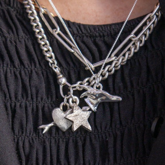 A close-up of a person wearing layered silver necklaces over a black ruched top. The necklaces include a chain necklace, a paperclip-style necklace, and a pendant necklace with a star, heart, and greyhound head charms.